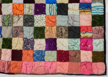 It features a grid of colorful squares stuffed with padding and scrunched together creating a very fluffy appearance.  The squares are made of a variety of fabrics, some plain and others with simple patterns. It is backed in silk that is embellished with silk embroidered flowers. A very unique and interesting piece.