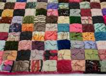 It features a grid of colorful squares stuffed with padding and scrunched together creating a very fluffy appearance.  The squares are made of a variety of fabrics, some plain and others with simple patterns. It is backed in silk that is embellished with silk embroidered flowers. A very unique and interesting piece.