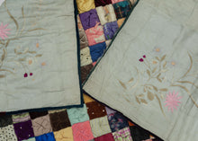 It features a grid of colorful squares stuffed with padding and scrunched together creating a very fluffy appearance.  The squares are made of a variety of fabrics, some plain and others with simple patterns. It is backed in silk that is embellished with silk embroidered flowers. A very unique and interesting piece.