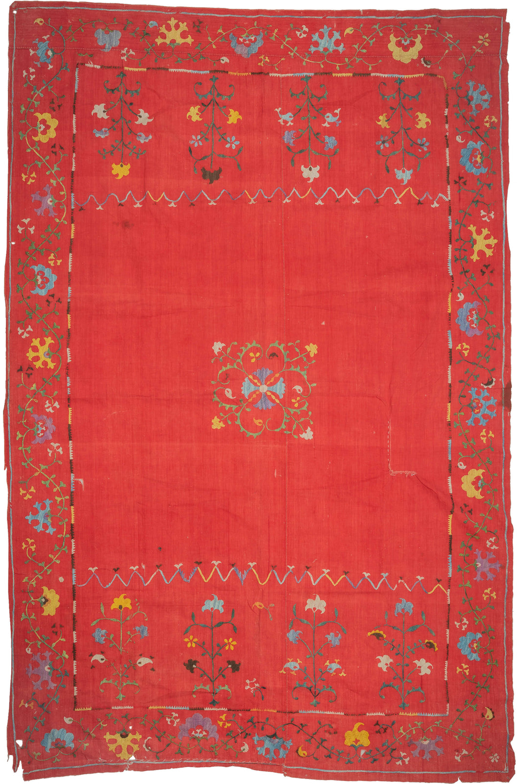 This Suzani was handmade during the early 20th century in Uzbekistan.
This suzani is composed of central floral motif embroidered in silk on an open red cotton field. Flanked by vibrant multi-tiered blossoming flowers to the right and left and framed by a lively floral meander. A multitude of colors are used for the embroidery, including turqoise, green, white, yellow, and purple.