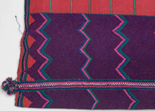A bold highland huipil woven with striking contrast of deep purple and brick red. The upper section is patterned with vivid zig-zags in pink, green, and turquoise, while the lower body panels feature a calmer striped red ground. Woven on a backstrap loom, this piece balances geometric energy with grounding simplicity, making it both a traditional garment and a statement textile.
