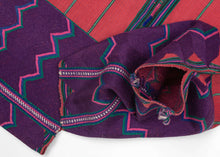 A bold highland huipil woven with striking contrast of deep purple and brick red. The upper section is patterned with vivid zig-zags in pink, green, and turquoise, while the lower body panels feature a calmer striped red ground. Woven on a backstrap loom, this piece balances geometric energy with grounding simplicity, making it both a traditional garment and a statement textile.