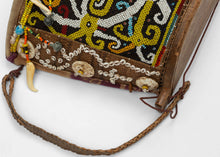 A richly adorned Dayak baby carrier from Borneo, made with a wooden frame, woven fiber body, resist-dyed cloth lining, and a densely beaded front panel. The design is worked in glass beads with bold yellow, white, black, red, and blue motifs, likely representing protective ancestral or spirit forms.

Among Dayak peoples, baby carriers were both practical and symbolic. Worn on the back, they held the child securely while also serving as objects of protection, status, and family care. Beadwork, shells, bone o