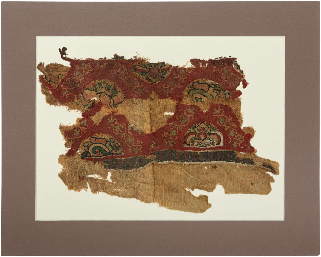 An exceptionally early Coptic textile fragment, woven more than 1,400 years ago, preserving richly patterned tapestry bands against a warm linen ground. Deep red fields are animated with scrolling vegetal forms and inset medallions containing highly stylized floral, animal, and possibly scarab-like motifs in ochre, green, cream, and dark brown.