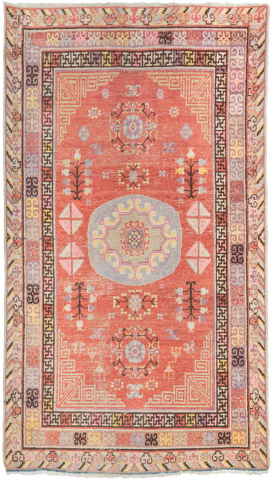This Khotan rug was woven in  central medallion in soft purples, pink, yellow and green atop a dramatic red field which also features diamonds and blossoming pomegranites. It showcases classic Khotan patterning which is a wonderful hybrid of both Central and Eastern Asian design elements like the lattice cornices, tri-color cloud band outer border, and greek-key interior border which latch hooked elements