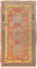 This Khotan rug was woven in Northwest China, which is also known as East Turkestan, during the early 20th century.   This rug features three orb-like medallions of atop a patinated orange-red ground of personality filed palmettes, rosettes and cloudbands. Thick solid color lines frame two main borders. One a wonky greek key on a blue ground in various states of fading and the other polychrome "shou" symbols on a stunning canary yellow ground. Shou is a very auspicious symbol which represents longevity.