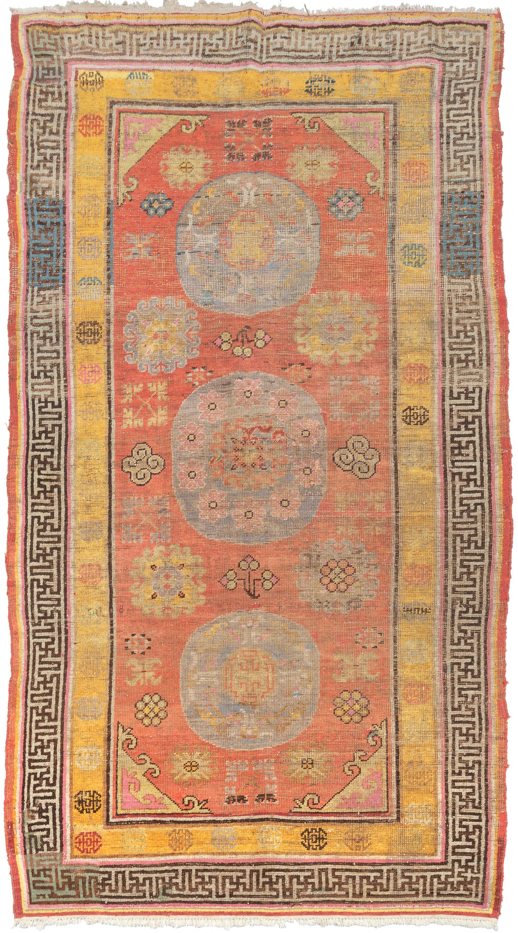 This Khotan rug was woven in Northwest China, which is also known as East Turkestan, during the early 20th century.   This rug features three orb-like medallions of atop a patinated orange-red ground of personality filed palmettes, rosettes and cloudbands. Thick solid color lines frame two main borders. One a wonky greek key on a blue ground in various states of fading and the other polychrome 