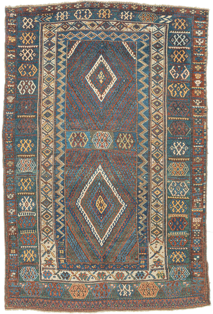 This rug was likely woven likely in the Hakkari - Rawanduz corridor of the Kurdish Highlands of Iraq during the turn of the 20th century

This deeply expressive Kurdish village rug presents a masterclass in tribal geometry, color harmony, and cultural symbolism. It features a field anchored by two nested diamond medallions, each filled with small, rhythmic glyph-like motifs and set against a ground of saturated indigo blue and madder red abrashed into glowing mineral shades. Surrounding the central field ar