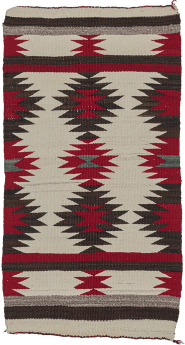 This Navajo rug was handwoven in the southwest United States in the middle of the 20th century.  It features a repeating sunburst pattern in brown, red, and gray on an ivory ground. The pattern features two horizontal bands of solid red and brown in the interior and is finished with solid horizontal bands of gray, brown, and ivory on both ends. 