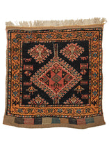 Small nomadic Qashqa'i bagface weaving depicting several figures including combs and two women around a diamond medallion. It features a warm color palette of oranges and reds on a dark field. 
