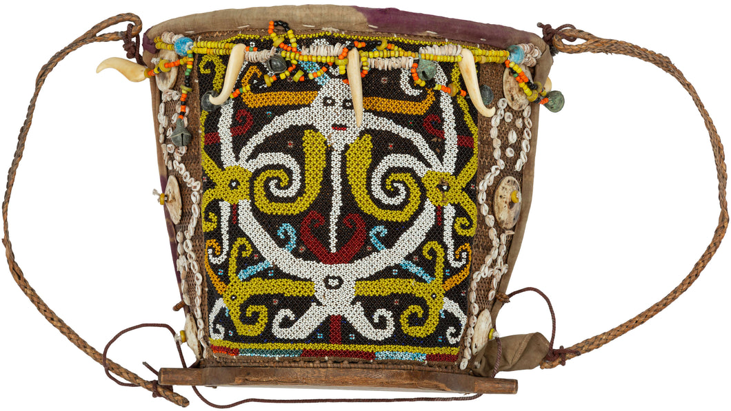 A richly adorned Dayak baby carrier from Borneo, made with a wooden frame, woven fiber body, resist-dyed cloth lining, and a densely beaded front panel. The design is worked in glass beads with bold yellow, white, black, red, and blue motifs, likely representing protective ancestral or spirit forms.

Among Dayak peoples, baby carriers were both practical and symbolic. Worn on the back, they held the child securely while also serving as objects of protection, status, and family care. Beadwork, shells, bone o