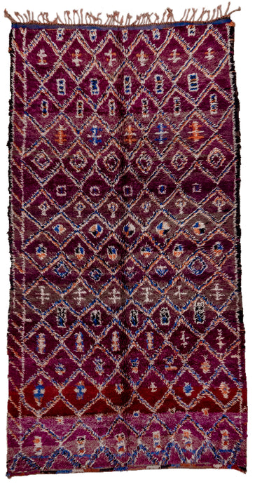 This Marmoucha rug was woven in the Middle Atlas mountains during the 20th century.  It features a diamond lattice design in orange, ivory and blue on a purple ground. Various purples are present shifting from wine to grape soda. The lattice is filled with simple shapes and protection symbols. There is a partial border only to one side. 