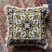 A vintage Portuguese decorative needlepoint pillow with a floral pattern and a geometric central medallion, fringed on all four sides, placed on a patterned rug.
