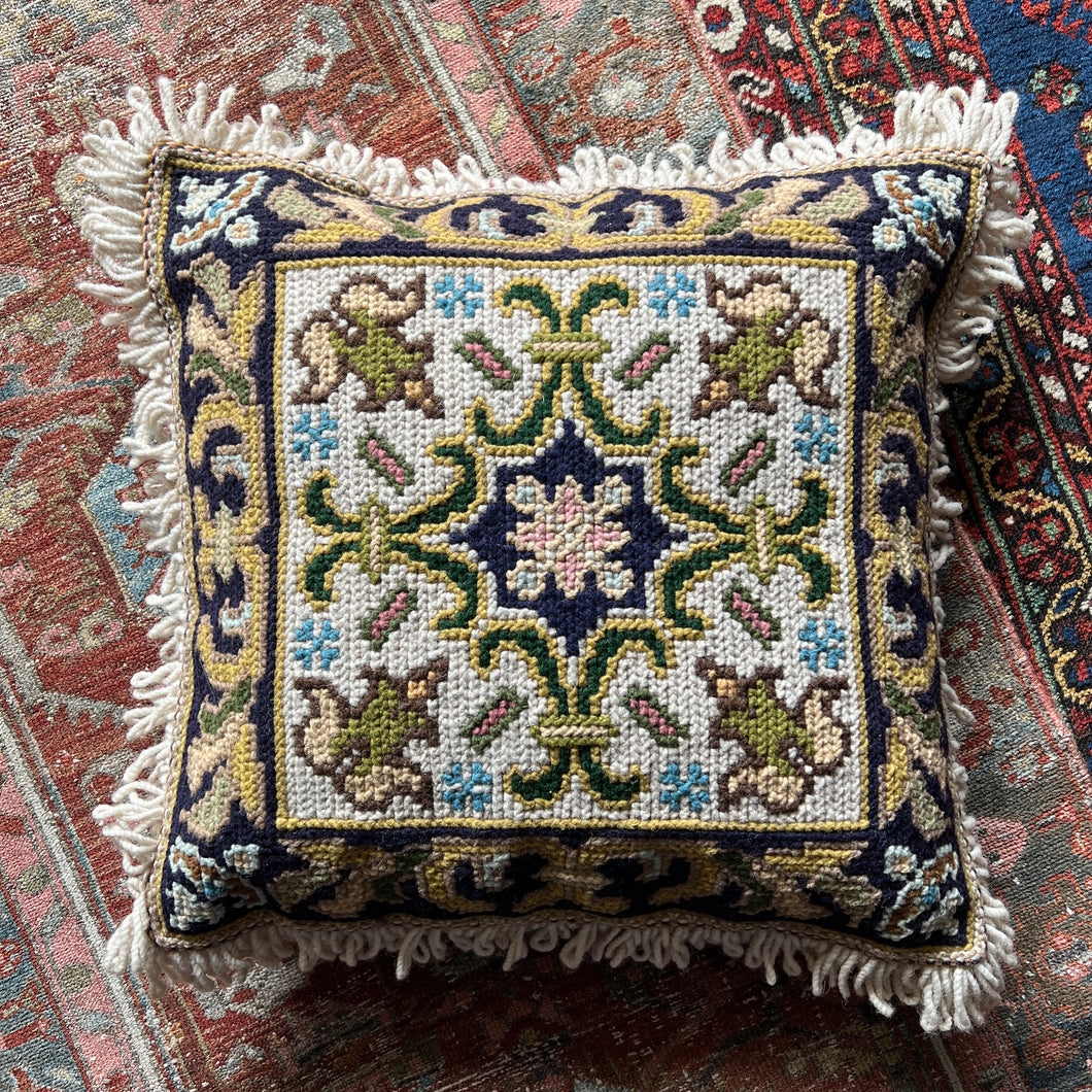 A vintage Portuguese decorative needlepoint pillow with a floral pattern and a geometric central medallion, fringed on all four sides, placed on a patterned rug.