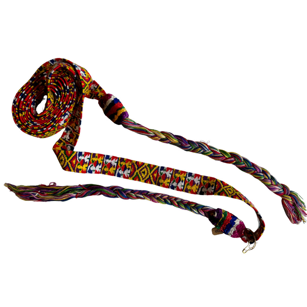 A vibrant woven cinta from Zunil, Quetzaltenango, Guatemala. This long headdress band is brocaded with repeating multicolored motifs, including geometric diamonds and stylized human figures in bright yellow, green, red, and white against a richly patterned ground. The ends finish in twisted multicolored tassels, typical of Zunil weaving.

Cintas are among the most distinctive elements of Maya women’s attire. They are wrapped elaborately around the head, creating a striking crown-like headdress, as seen in t