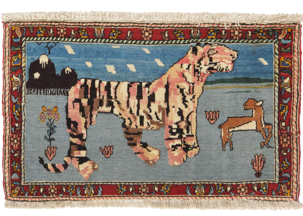 vintage Bakhtiari Pictoral rug featuring a large tiger on a blue field with a small deer to the right. In the background are mountains with clouds and birds in the sky. In the foreground of the scene are flowers and grass.