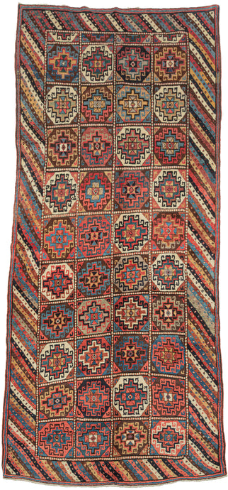 A boldly patterned antique Kurdish long rug woven with a striking compartment design. The field is organized into a grid of stepped, octagonal Memling guls, each enclosing a central rosette, rendered in a rich palette of madder red, indigo blue, ivory, soft camel, brown, and green.

The Memling gul, an ancient geometric motif appearing across many Caucasian and Kurdish weaving traditions, gives the rug its powerful visual structure. Here, the repeated guls are framed by an especially lively diagonal-striped