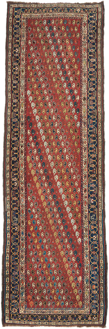 A&nbsp;madder red field densely packed with an all-over diagonal boteh repeat in ivory, navy, gold, teal, and brown forms scattered across the ground in a loose, lively arrangement. The runner reads almost like a textile print at distance and reveals real drawing up close. No two botehs are identical; the color varies row to row, giving the field a warm, animated quality that a more regimented repeat couldn’t achieve.

The main border is a deep navy with a bold reciprocal leaf-and-vine pattern, framed by na