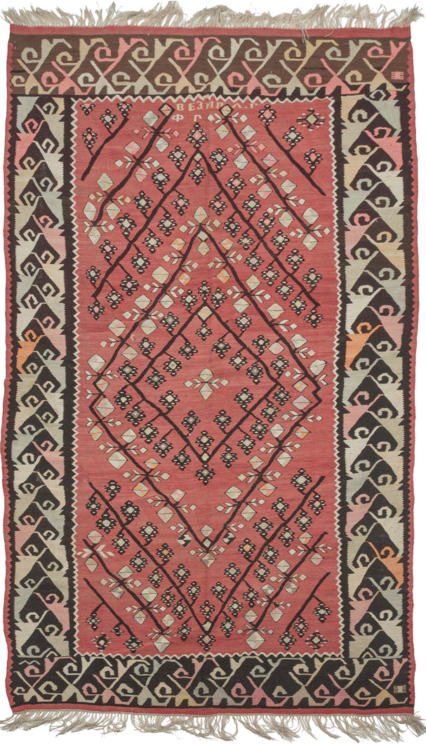 This bold and finely executed flatwoven kilim from Pirot, Serbia, features a rhythmic lattice of diamonds and blossoming bud motifs arranged across a vibrant red field. The powerful graphic border is composed of stylized, angular vine-scroll forms and rendered in tones of black, ivory, ochre, pink, and apricot.

At the top, the woven Cyrillic inscription “ВЕЗИРАR” (Bezirac) suggests a prestigious workshop or district, likely tied to a“Bezir” (vizier) a high-ranking Ottoman official. This connection reflects