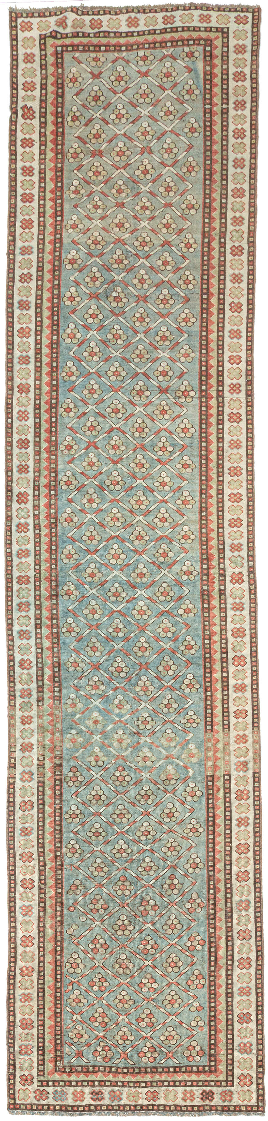 This long Kurdish runner features a softly faded, open lattice of floral rosettes set within a repeating diamond framework, giving the piece a gentle rhythm well suited to its elongated format. The field’s washed out pale blue-green ground has a restrained, almost pastel register that feels calm and architectural rather than decorative.