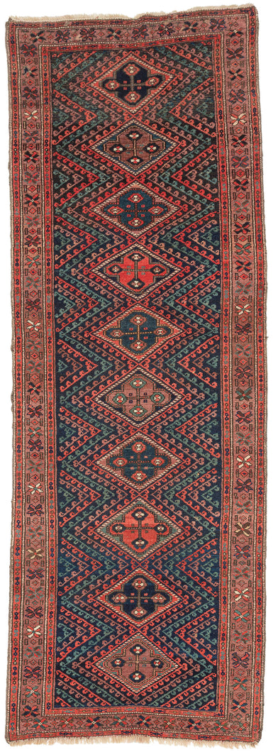A richly colored antique Kurdish runner with a dramatic dark field and a vertical sequence of stepped diamond medallions. Each medallion contains a bold four-part floral form, set within a field of angular zigzag bands and small hooked motifs in madder red, deep indigo, soft rose, aubergine, and green.

The rug has a strong tribal character, with a narrow format, saturated color, and confident geometric drawing. The repeated diamond medallions create a clear central rhythm, while the surrounding serrated fo