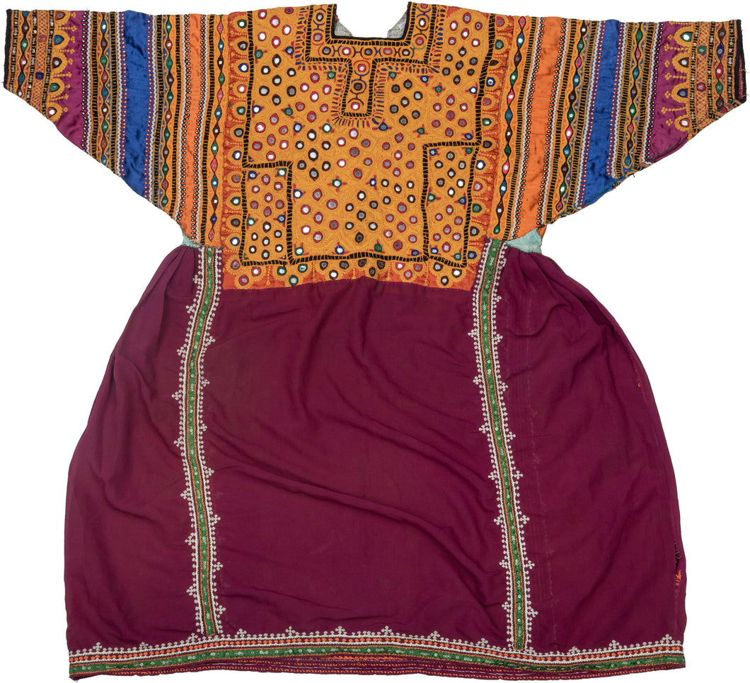 This striking vintage dress from Sindh, Pakistan, showcases the region’s renowned textile artistry through dazzling mirrorwork embroidery (shisha) on a richly dyed base. The bodice and sleeves are densely adorned with geometric mirror insets, chain-stitch embroidery, and floral motifs, worked in bold jewel tones on ochre, orange, and cobalt panels. The lower portion of the dress is a deep maroon cotton, finely detailed with vertical bands of white bead-like embroidery and green piping, adding balance and el