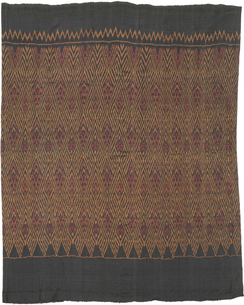 This elegant silk pha sin or traditional tube skirt was handmade northeast Thailand during the middle of the 20th century.

It is a masterful example of mudmee (ikat) weaving from the Isaan region. Woven entirely from fine silk, the textile features a mesmerizing field of diamond motifs arranged in vertical zigzag bands, subtly interlaced with red, gold, and deep aubergine tones against a near-black ground. The effect is one of shimmering depth and rhythmic precision.

The upper and lower bands (hua sinh an