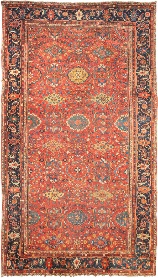 It features a dense and rhythmic allover pattern of palmettes, rosettes, and roving vinery set against a richly saturated madder red field. Lush floral clusters and shield-like medallions in blues, golds, ivory, and greens are symmetrically arrayed across the surface, producing a mesmerizing, layered composition. The deep indigo border is animated with bold palmettes and angular vine scrolls, grounding the exuberant interior with contrast and balance. This carpet’s large format and harmonious pale
