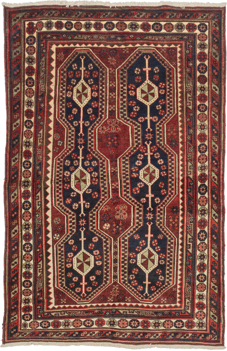 A Richly colored tribal rug with a striking double-column format of two tall elongated medallions&nbsp;on a deep indigo ground, each structured around a vertical spine of diamond forms quartered in olive and cream. The m&nbsp;red field between and around them reads almost as a third element, giving the composition an unusual density. Colors throughout are well-preserved with navy, red, ivory, and a muted green that surfaces in the medallion interiors and guard borders.