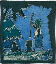 A striking West African batik textile worked in deep indigo, teal, and blue tones. The composition shows a standing figure using a tall pestle with a mortar, set within a stylized village landscape with a thatched structure, bowls, palms, and birds.