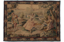 Antique  French Aubusson tapestry depicting a bucolic scene of a "coquette" and two potential suitors. At the center of the scene, a woman lifts her skirts to reveal an ankle towards a man propped up against a tree, playing a lute. A second man passes by and both him and the woman take notice of another. A house is visible in the background, as is a body of water. A very dainty meander of ribbon and flowers frames the scene as the border. 