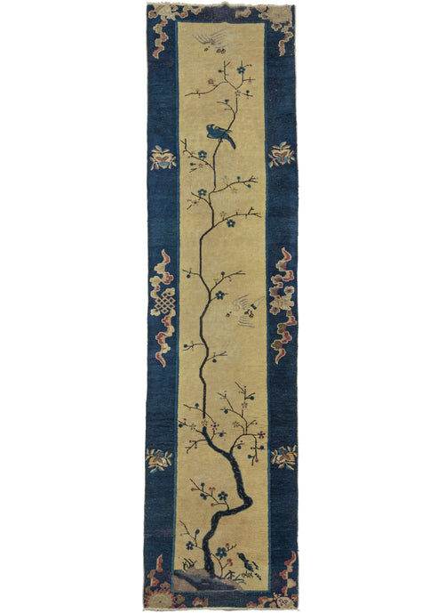 This Tree Deco Runner Rug features a thin tree growing upwards in a very organic manner on a yellow ground. The branches are sparsely adorned with buds and blossoming rosettes. A blue bird is perched on one of the branches near the top as one yellow bird flies above it and another yellow bird flies below. It is framed by a navy border that is mostly open except for various auspicious symbols wrapped in bent ribbon cloud bands.