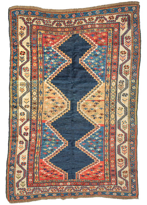 This Antique Lori Rug features a connected column of lively stepped diamonds rendered in a handsome navy. The solid navy column is unadorned and stands out against a backdrop containing big blocks of vibrant yellow, red and softer indigo that contain abstracted boteh or bird-like figures. The main border features a bright ivory ground and scrolling vinework which alternates with directional blossoming flowers along the sides and more compact rosettes on the top and bottom.