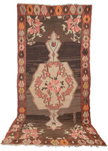 Turkish Kars Kilim featuring a scalloped central medallion with a dainty floral center surrounded by pink scrolls on camel ground. The same delicate flower featured in the medallion appears in each cornice. The field features a lovely brown which undulates wildly from light to dark brown which gives a subtle but pulsating energy. The word "BEHREM" which may be the weaver's name has been woven into the bottom right quadrant. The whole is framed by an abstracted floral motif. 