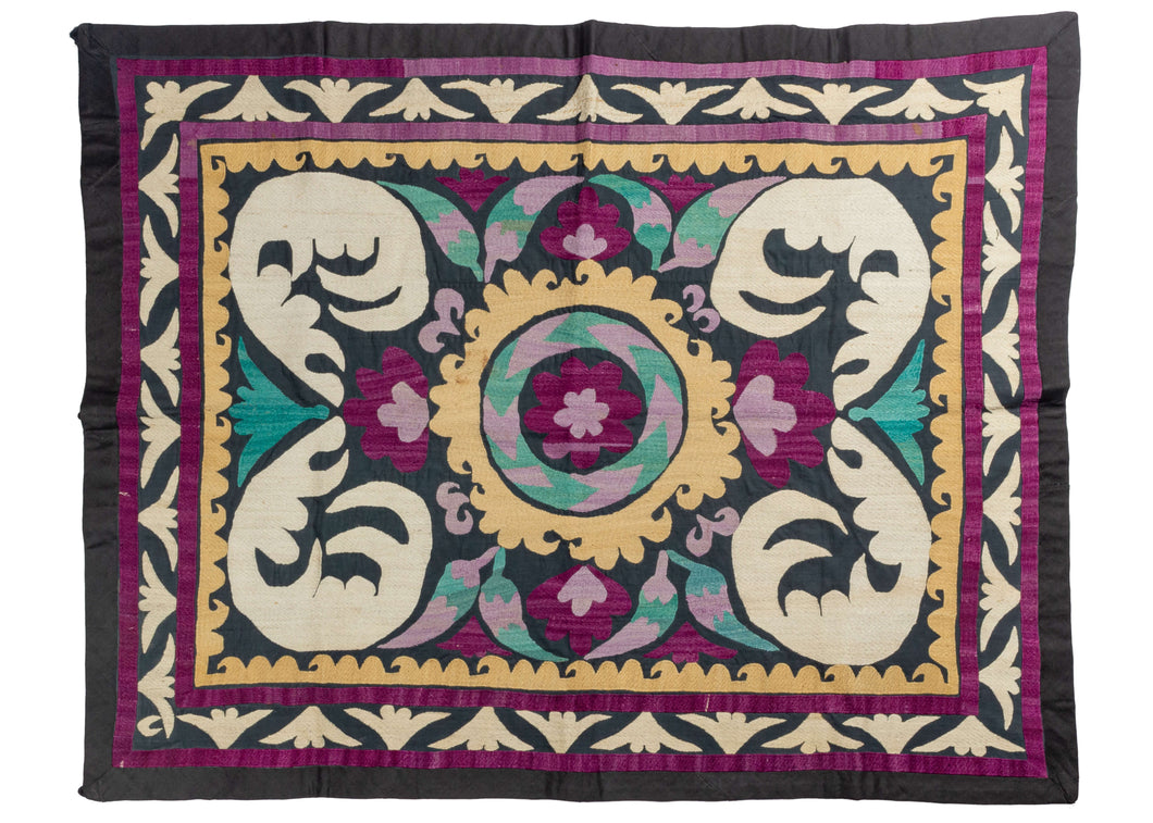 Central Asian Uzbek Suzani wall hanging featuring a circular central medallion with two-tone purple rosettes in the center and on each side. Curled white leaves that are reminiscent of a floating feather can be found in each corner. Beautiful accent tones of gold and emerald add extra interest and really pop with the other tones against the black field.