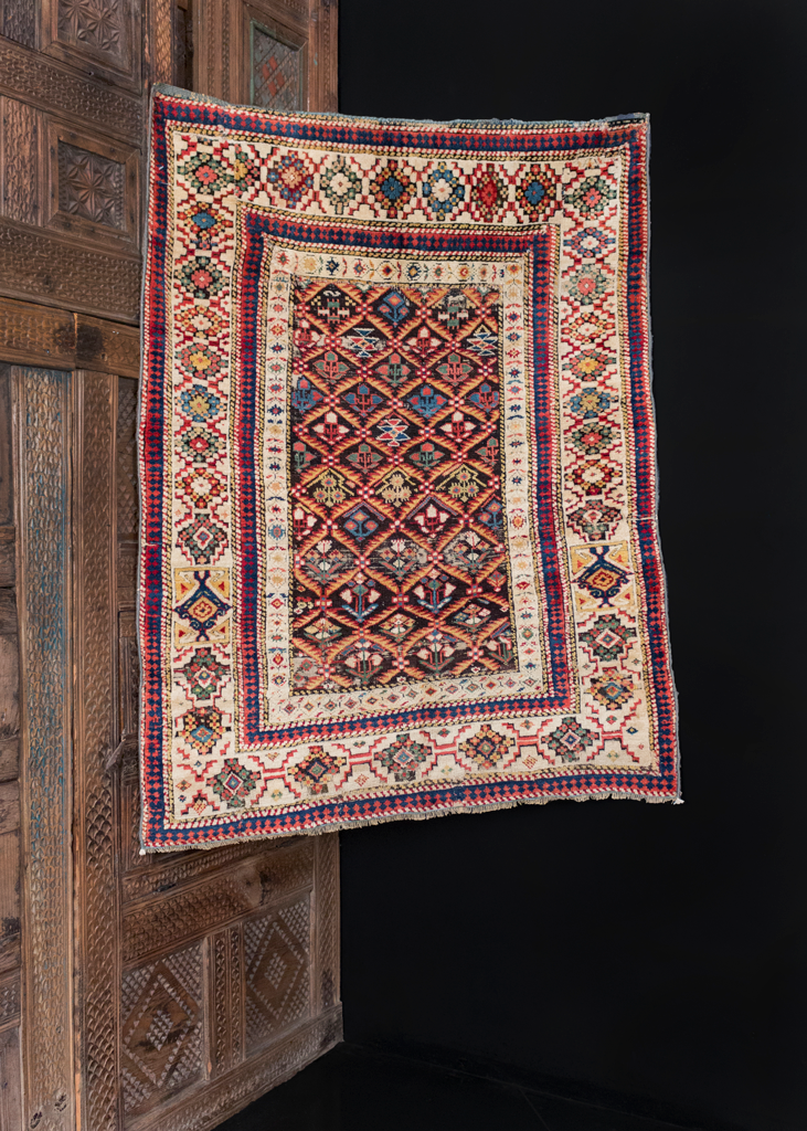 Shirvan rug handwoven during middle of 19th century in the Caucasus. Colorful array of blossoming flowers separated by yellow lattice atop dark field. Large ivory border surrounds field with brightly colored bouquets. 
