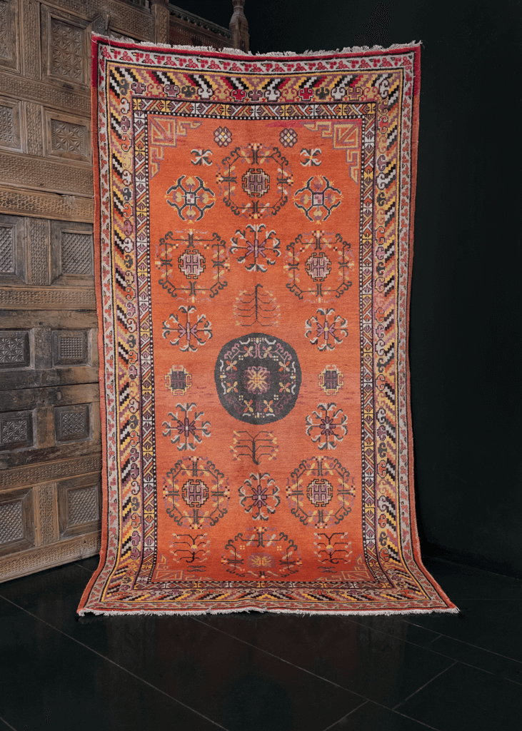 Khotan rug handwoven during second quarter of 20th century in Northwest China. Grey medallion with geometric floral drawings atop orange field. Showcases East Asian design elements like the lattice cornices and cloud band border. 