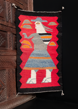 Navajo pictorial rug with a red field and black border. A woman is woven wearing a hat with a sack on her back. 