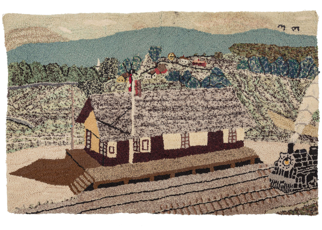 Thus Vintage Train Station Hooked Rug features an idyllic scene of a black locomotive pulling into a station. The station is shown in the foreground with a small village behind it in a lush, hilly landscape. Speckled yarn is used in the station's roof as well as the landscape behind adding some layers while the smoke billowing over the incoming train and the shadow to the left of the station add movement and life to the composition. 
