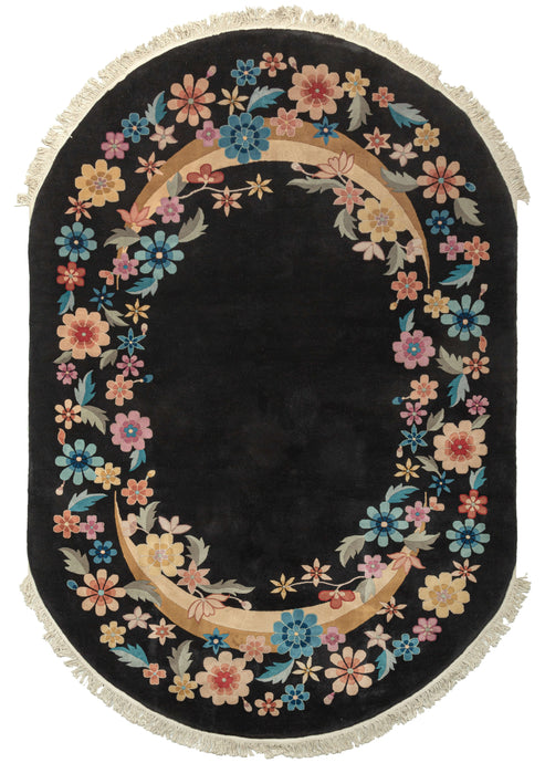 Plush black oval Chinese deco area rug featuring a true black field, with an oval ring of various multicolored flowers. Blue, pink, orange, and yellow blossoms can be identified as stylized peonies, narcissus, chrysanthemum, lotus, plum blossoms, and peach blossoms. The cut-pile technique of cutting piles at different angles to sculpt and shape the wool is used to stunning effect in this rug. 