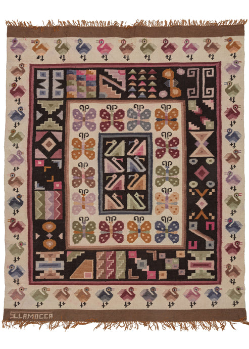 This vintage Signed Peruvian Kilim It features a central square of polychrome swan-like birds on a black ground surrounded by a series of concentric borders. They feature a band of butterflies on a white ground, various one-off geometric symbols on a black ground, and finally a border of birds of a different variety than what was utilized in the center. The top and bottom are finished with undyed brown skirt borders. The bottom skirt is signed by the weaver 