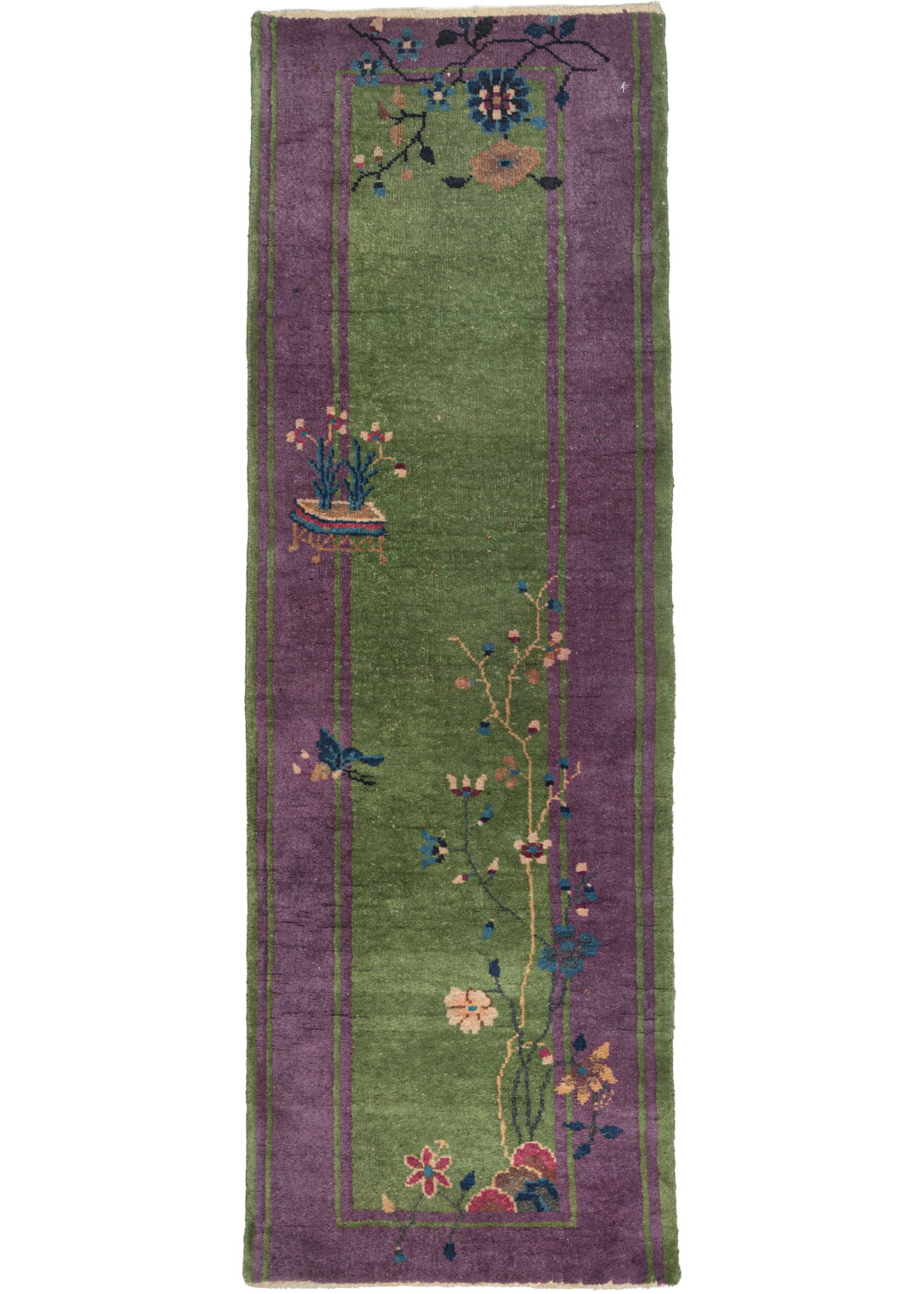 This Purple and Green Deco Runner features a striking and desirable contrast of a rich green ground encapsulated by a regal purple border. Various vegetation including a small flowering tree and flowers in a plant holder pays no mind to the barrier between border and field giving this piece a feeling that is free and unrestrained. In a hard-to-find short and narrow runner format.