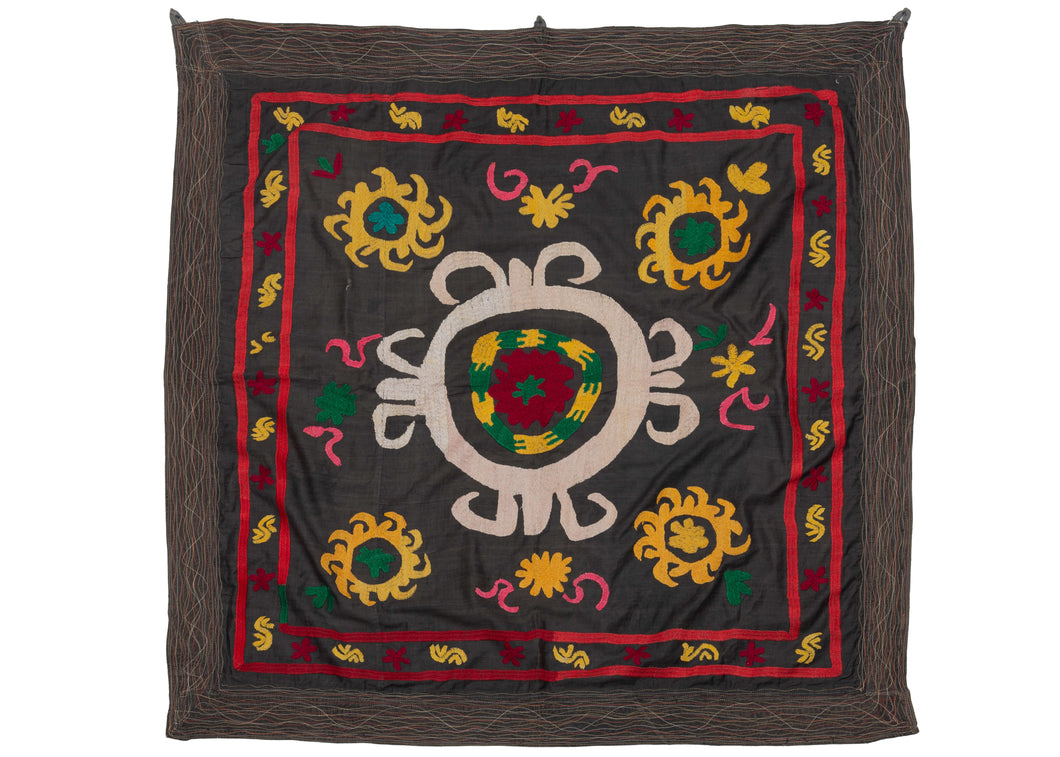 This Vintage Uzbek Suzani features a central medallion in white, red, yellow, and green atop a faded black ground. Four smaller medallions in yellow and green are in each corner of the piece and the whole is framed by a border. The handmade quality of this textile is apparent in the asymmetrical nature of the embroidery giving the medallions movement and distinct personality.