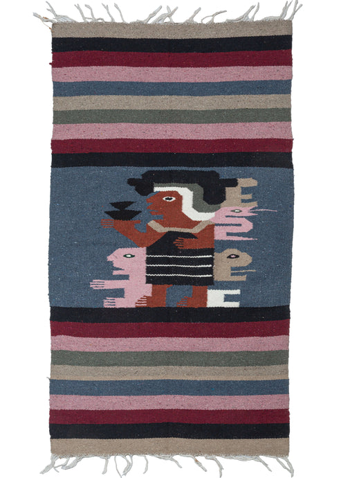 This Vintage Mexican Tapestry features a central design of a copper-toned figure in profile wearing black and white Mayan attire and holding a cup of what may be a Cocoa drink. A variety of animal forms are displayed around the figure in soft pink and a sandy brown.