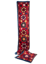 This Vintage Uzbek Suzani Welcome Banner features a row of large rosettes which are constructed of smaller rosettes in blue, yellow, white, and navy on a bright red field. A black stitched border and blue fringe can be found surrounding its entire perimeter.