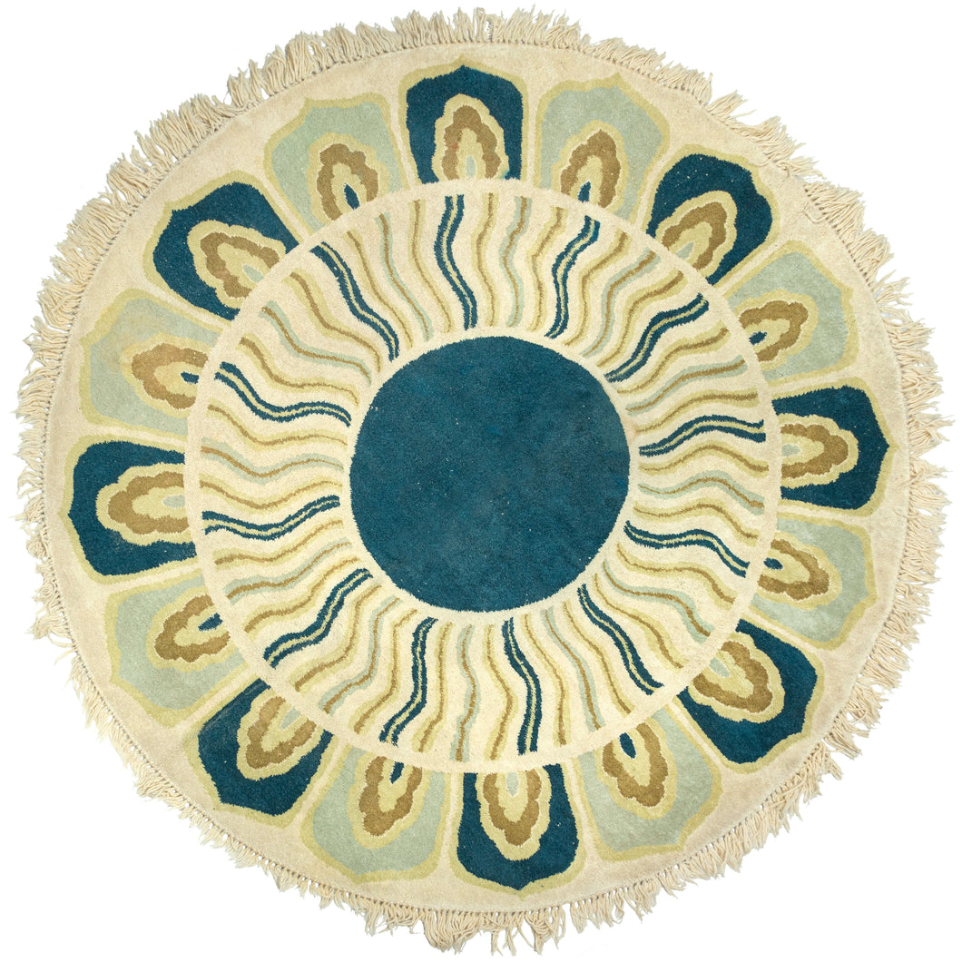 This Mid Century Indian Round Rug features a simple color palette of ivory, sage, gold, and a rich blue/green. A solid central circle is encapsulated by a second circle of wavy lanes and a final circle of petal-like ornamentation. The rug has a very mod/swinging 60s feel with a design reminiscent of a psychedelic sun or giant rosette. Finished with a fringe surrounding the whole perimeter. 